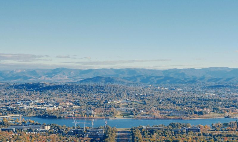 A breathtaking aerial view of Canberra city with mountains and vibrant autumn foliage under a clear blue sky.